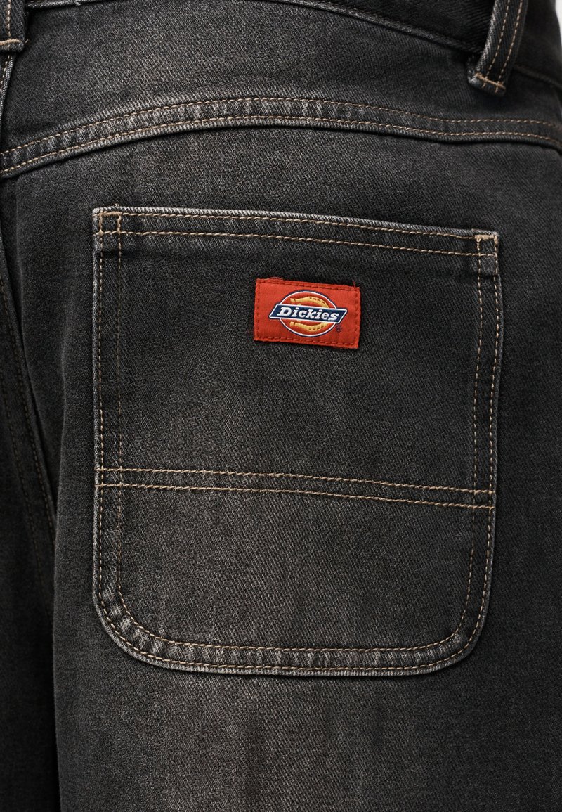Gesäßtasche einer schwarzen Denim-Hose mit sichtbaren Nähten und einem roten rechteckigen Dickies Markenlabel, das über der Tasche aufgenäht ist.