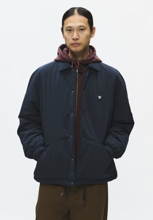 Jeune homme aux longs cheveux attachés, portant une veste bleu marine par-dessus un sweat à capuche bordeaux et un pantalon marron, debout les mains dans les poches sur fond blanc.