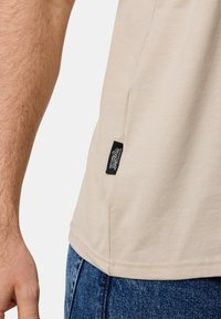 Beige t-paita, jossa on pehmeä pinta, mustalla brändilapulla sivusaumassa ja lyhyet hihat. Yhdistettynä sinisiin farkkuihin.