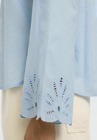 Helehallide pika varrukaga särk, millel on keerukas pitskudumine varrukalõpus ja laineline äär. Pehme kangasteksts.