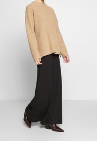 Maglione beige a coste con una vestibilità rilassata, scollatura rotonda e spalle cadenti, abbinato a pantaloni neri a gamba larga e stivali con tacco.