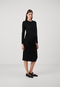 Vestido de maternidade preto de mangas longas, justo com lados franzidos, até ao joelho, de textura de tecido suave, combinado com sapatos pretos planos.