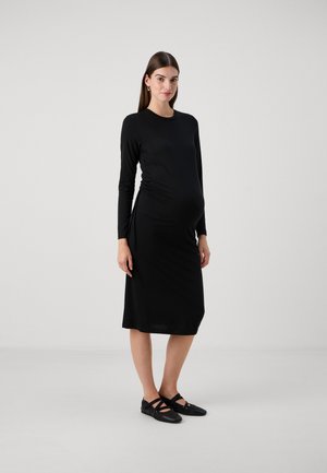 Robe de maternité noire à manches longues, ajustée avec des côtés froncés, longueur genou, texture de tissu douce, associée à des chaussures plates noires.
