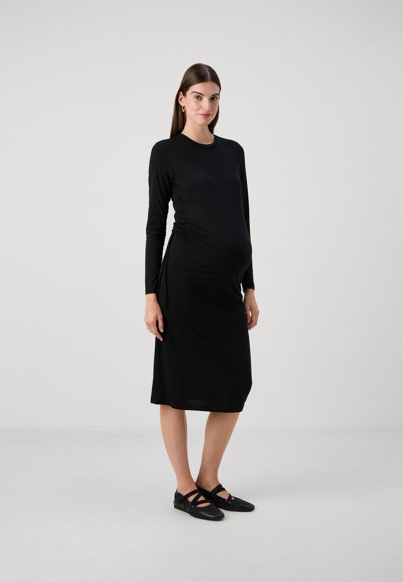 Vestido de maternidade preto de mangas longas, justo com lados franzidos, até ao joelho, de textura de tecido suave, combinado com sapatos pretos planos.