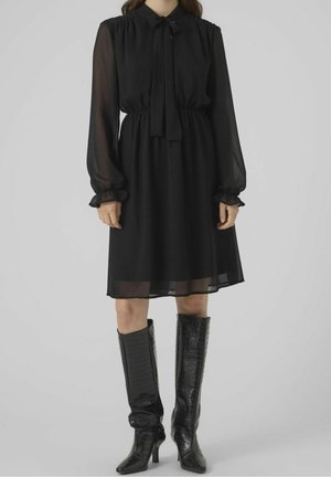 Femme portant une robe noire à manches longues avec des manches transparentes et des bottes hautes noires jusqu'aux genoux avec un motif texturé.