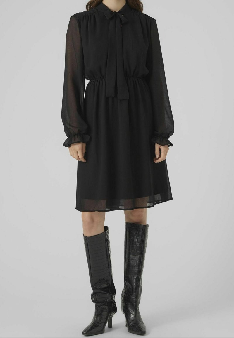 Femme portant une robe noire à manches longues avec des manches transparentes et des bottes hautes noires jusqu'aux genoux avec un motif texturé.