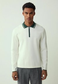 Man wearing white long-sleeve polo shirt with dark green collar and gray trousers, standing against plain light background.