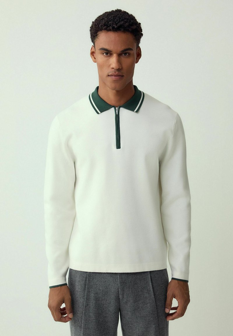 Man wearing white long-sleeve polo shirt with dark green collar and gray trousers, standing against plain light background.