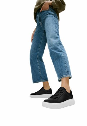 Persoon draagt omgeslagen blauwe spijkerbroek en zwarte sneakers met dikke witte zolen, staande tegen een witte achtergrond.