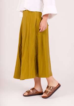 Person trägt einen senffarbenen Midi-Rock mit weißer Bluse und braunen Slip-On-Sandalen mit zwei Schnallen und lasergeschnittenen Details.