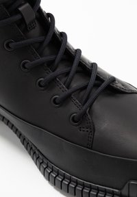 Bota de cuero negra con punta redonda, suela de goma plana y cordones negros. Presenta ojales de metal y detalles de costura en el lateral.