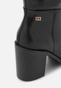 Bottine en cuir noir avec un talon large, une texture lisse et un petit emblème doré sur le côté. Des coutures décoratives soulignent le design.