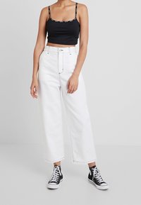 Kvinna iklädd en svart croptop med logoband, högmidjade vida vita byxor och svarta högskurna sneakers, som står mot en vit bakgrund.