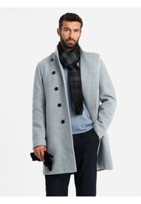Cappotto in lana grigio chiaro con grandi bottoni neri, colletto alto e tasche laterali, abbinato a una sciarpa a quadri scuri e guanti neri.