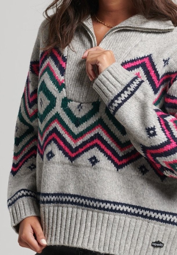 Femme portant un pull en maille gris avec des motifs en zigzag et en diamant rose, vert et bleu marine, doté d'un col à fermeture éclair quart de tour et de poignets côtelés.