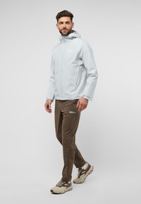 Lichtgrijze waterdichte jas met capuchon, gecombineerd met bruine, taps toelopende broeken. Schoenen zijn beige met bruine accenten en een gestructureerde zool.