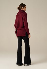 Donna con i capelli castani raccolti in un bun, che indossa un maglione spesso bordeaux, pantaloni neri a zampa d'elefante e tacchi a punta neri, in posizione laterale.