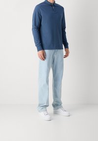 Koszula polo z długim rękawem w ciemnoniebieskim kolorze, z kołnierzykiem i zapięciem na guziki, w połączeniu z jasnoniebieskimi dżinsami i białymi trampkami. Prosta, gładka faktura.