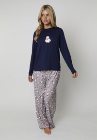 Donna che indossa una maglietta a maniche lunghe blu navy con una grafica di pinguino e pantaloni da pigiama grigi con motivi di pinguini e fiocchi di neve rossi e bianchi.