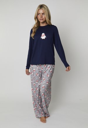 Vrouw in een marineblauwe longsleeve top met een pinguïnprint en een grijze pyjamabroek met rood-witte pinguïn- en sneeuwvlokpatronen.