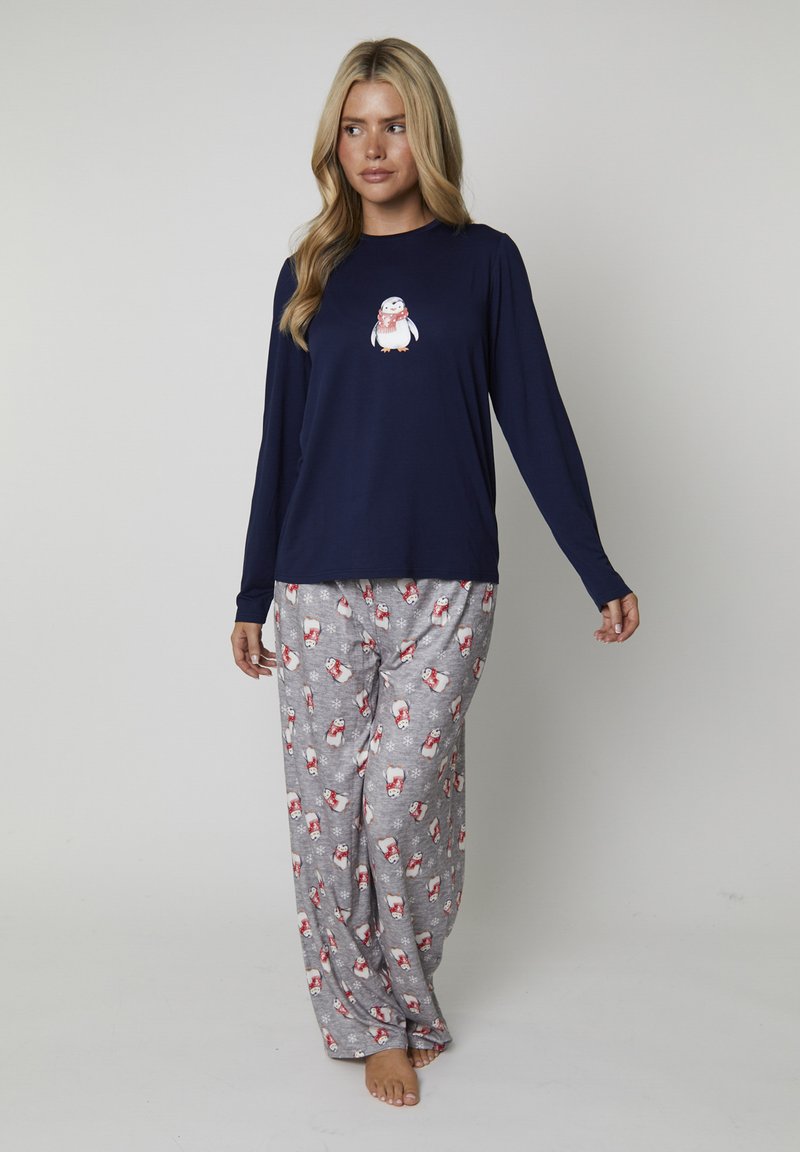 Donna che indossa una maglietta a maniche lunghe blu navy con una grafica di pinguino e pantaloni da pigiama grigi con motivi di pinguini e fiocchi di neve rossi e bianchi.