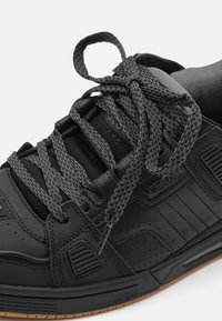 Black sneaker with a textured leather upper, grey laces, padded tongue, and a gum rubber sole. Features branding on the side.