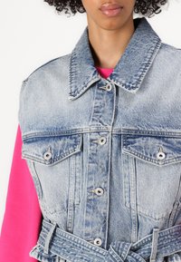 Gilet di denim azzurro con colletto, bottoni a pressione, due tasche frontali e vita con cintura; indossato sopra un indumento rosa.