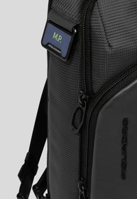 Mochila de tela negra con superficie texturizada, que presenta una etiqueta de nombre rectangular, bolsillos con cremallera y un detalle de cuero con logo en relieve.