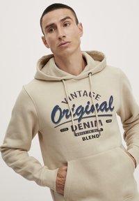 Sudadera con capucha beige con un bolsillo frontal, adornada con un estampado gráfico en azul marino que dice "Vintage Original Denim Blend". Material suave y puños acanalados.