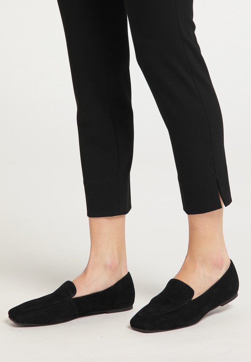 Zwarte suède instappers met een puntige neus, platte zool en een slip-on ontwerp, gecombineerd met op maat gemaakte zwarte broek met zijsplitten.