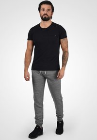Camiseta negra de manga corta con un bolsillo en el pecho, combinada con pantalones de chándal grises con cintura elástica. Zapatillas deportivas negras completan el look.
