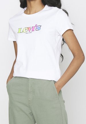 Młoda kobieta w białej koszulce Levi's z kolorowym logo i jasnooliwkowymi spodnimi, ręce w kieszeniach, na jednolitym tle.