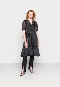 Vestido preto de cetim com mangas bufantes, decote em V e bainha com franzidos. Combinado com calças pretas ajustadas com fendas laterais e sandálias pretas.