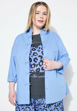 Camicia azzurro chiaro con taglio ampio, dotata di lacci decorativi, indossata sopra una camicia a motivo leopardato nero e blu.