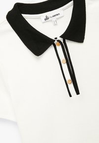 Hvid polo t-shirt med en sort ribbede krave, som har tre træknapper og lodrette sort-hvide striber langs stolpen.