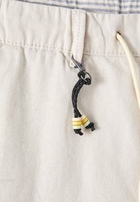 Tissu beige à texture chevron, équipé d'une fermeture à bascule avec un cordon noir tressé et des perles colorées. Détail de cordon visible.