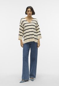 Maglione beige a righe con scollatura a V profonda, maniche ampie; abbinato a jeans blu a vita alta a zampa d'elefante e scarpe a punta di un blu chiaro.