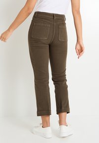 Pantalons en denim marron, coupés, avec une taille haute et deux poches arrière, associés à des baskets blanches. Le tissu semble lisse avec une texture minimale.