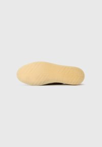 Skoens såle har en teksturert beige gummi med spor og merker for godt grep. Designet er flatt og strømlinjeformet.