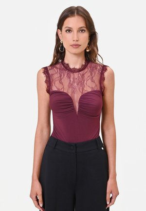 Donna che indossa un top bordeaux senza maniche con dettagli in pizzo e pantaloni neri a vita alta, in piedi davanti a uno sfondo semplice.