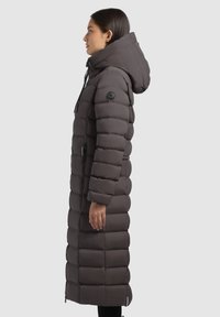 Cappotto lungo, imbottito, grigio antracite con cappuccio, dotato di chiusura con zip e design trapuntato. Accentuato da una toppa con logo sulla manica.