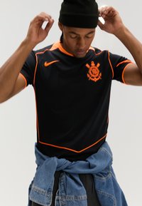 Svart atletisk trøye med oransje detaljer, med en logo og korte ermer. Brukes med en denimjakke knyttet rundt midjen.