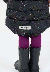 Barn iført sorte gummistøvler, lilla leggings og en sort puffjakke med farverige prikker og et "Zigzag" logo.