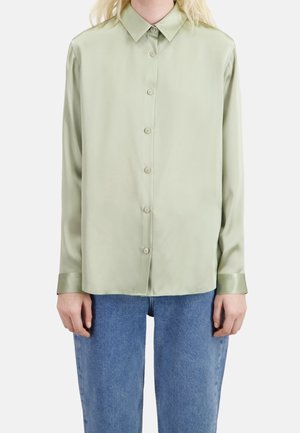 Person iført en lysegrøn skjorte med knapper og lange ærmer, kombineret med blå denimjeans, stående mod en ensfarvet lys baggrund.