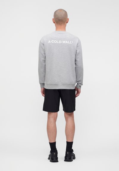 Άνδρας που στέκεται με την πλάτη, φορά γκρι φούτερ με το κείμενο «A-COLD-WALL*» στην πλάτη, μαύρα σορτς, μαύρες κάλτσες και μαύρα αθλητικά παπούτσια.