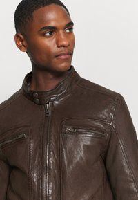 Giacca di pelle marrone con colletto alto, zip frontale e due tasche sul petto con zip. La texture è liscia con una leggera lucentezza.