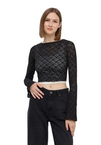 Top corto in pizzo nero con maniche lunghe, motivo floreale e scollatura tondeggiante; indossato sopra jeans neri a vita alta.