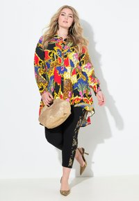 Blusa de colores con estampado, de manga larga, con dobladillo asimétrico y pantalones negros con detalles florales dorados, combinados con un bolso dorado y tacones.