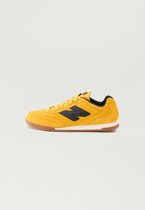 Yellow suede sneaker featuring a black logo, lace-up design, textured upper, and contrasting rubber sole with a gum-colored bottom.