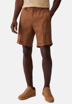 Uomo che indossa pantaloncini marroni con risvolti, camicia beige e sneakers in camoscio marrone abbinati, in piedi davanti a uno sfondo chiaro e uniforme.